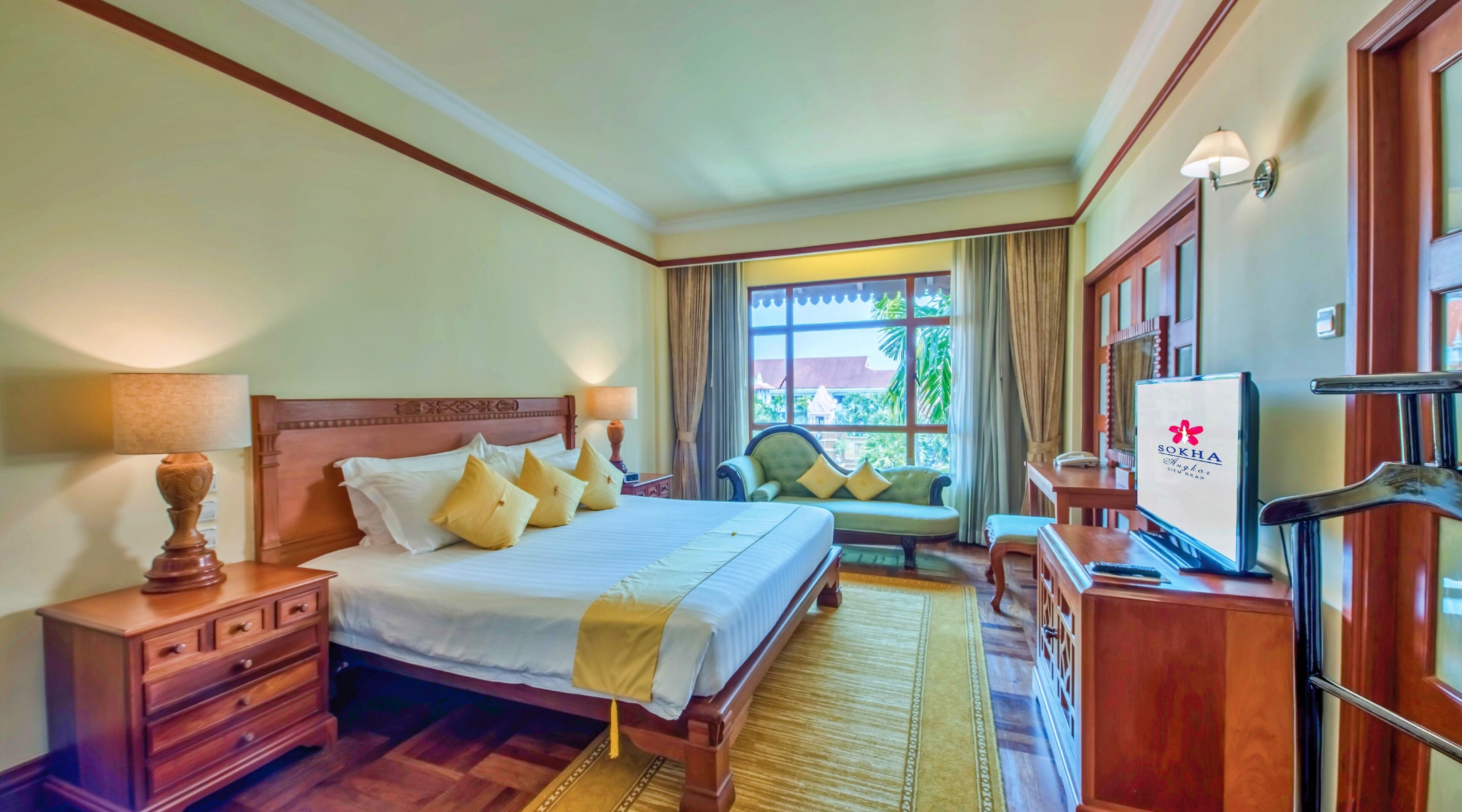 Quarto de hotel com cama, mesas de cabeceira, sofá, televisão e janela com cortinas.