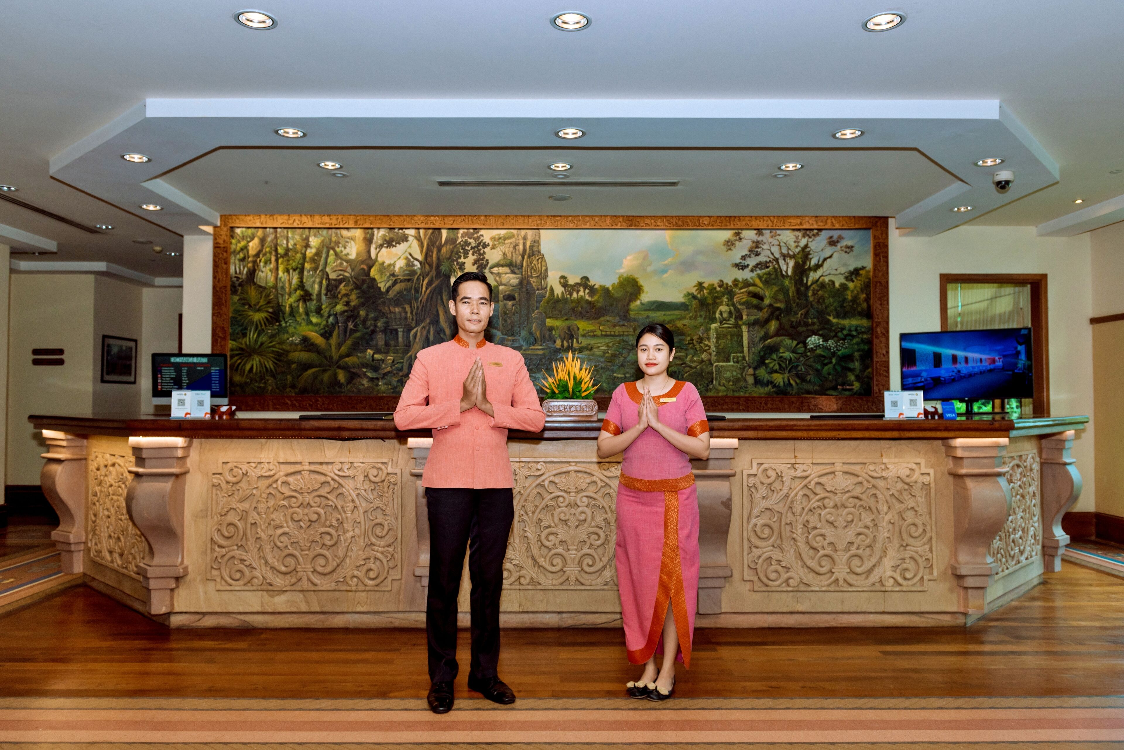 A reception desk with two individuals standing in front, a painting of a tropical landscape, and a television screen displaying a train.