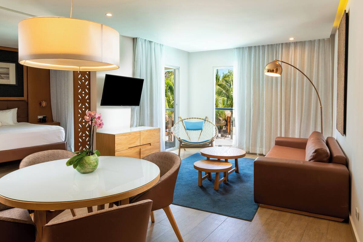 A modern hotel room with a bed, a dining area, a sofa, a TV, and a balcony with curtains.