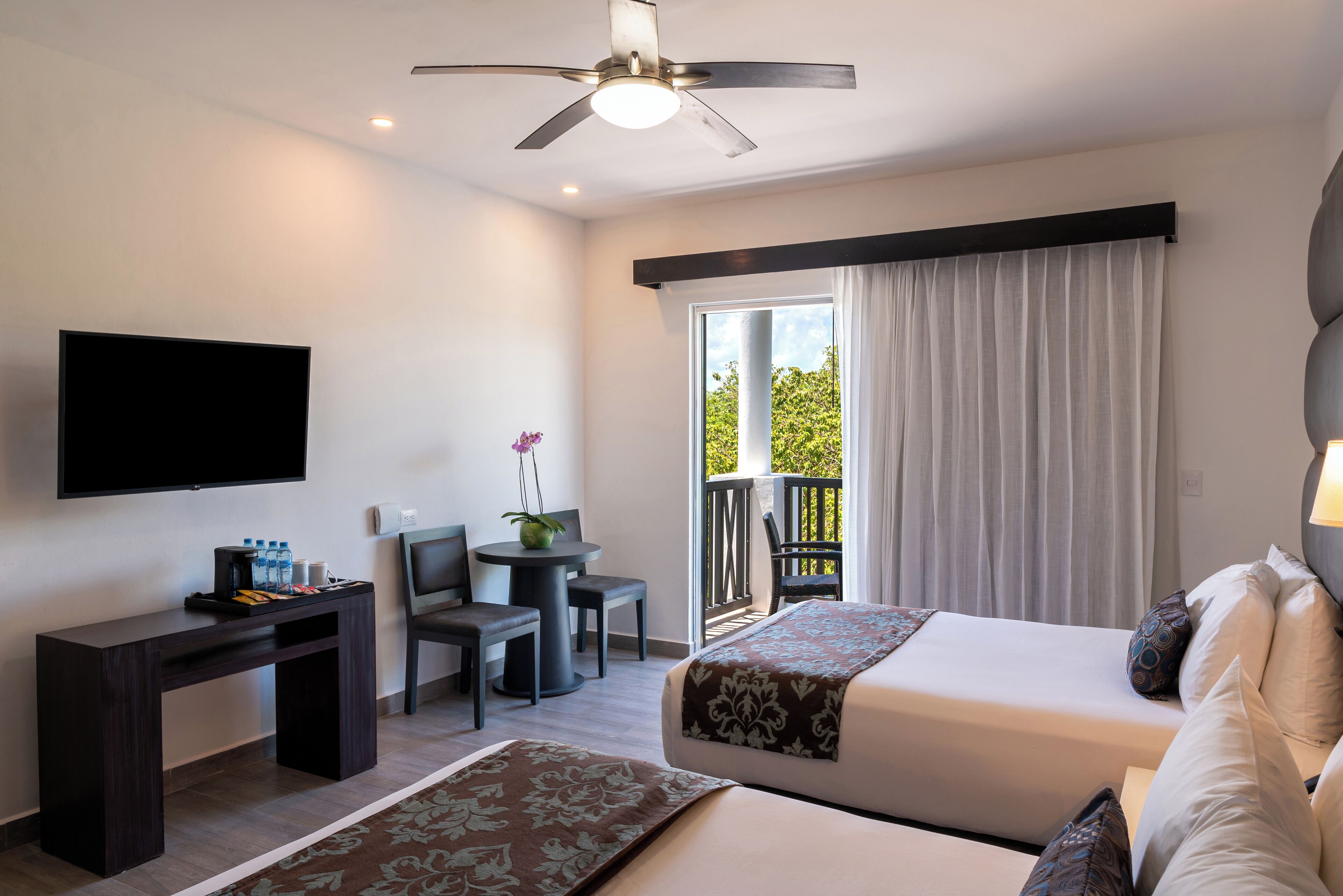 Une chambre d’hôtel comprenant un lit, un bureau avec un téléviseur à écran plat, une chaise, une petite table ornée d’un vase de fleurs et un balcon avec vue sur la verdure.