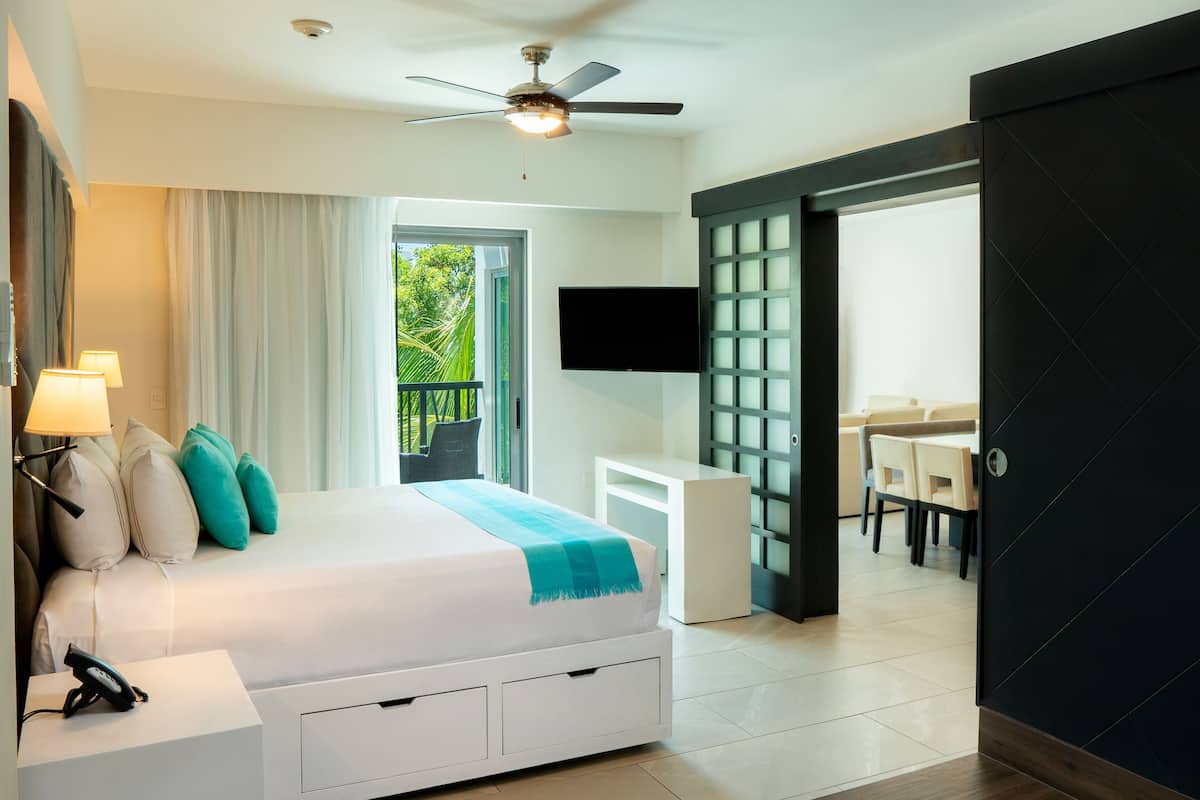 A modern hotel room with a bed, bedside lamps, a ceiling fan, a TV, and a balcony with a view of greenery.