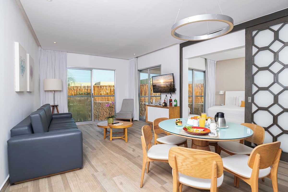 A modern hotel room with a sofa, dining table, chairs, a TV, and a balcony with a view.