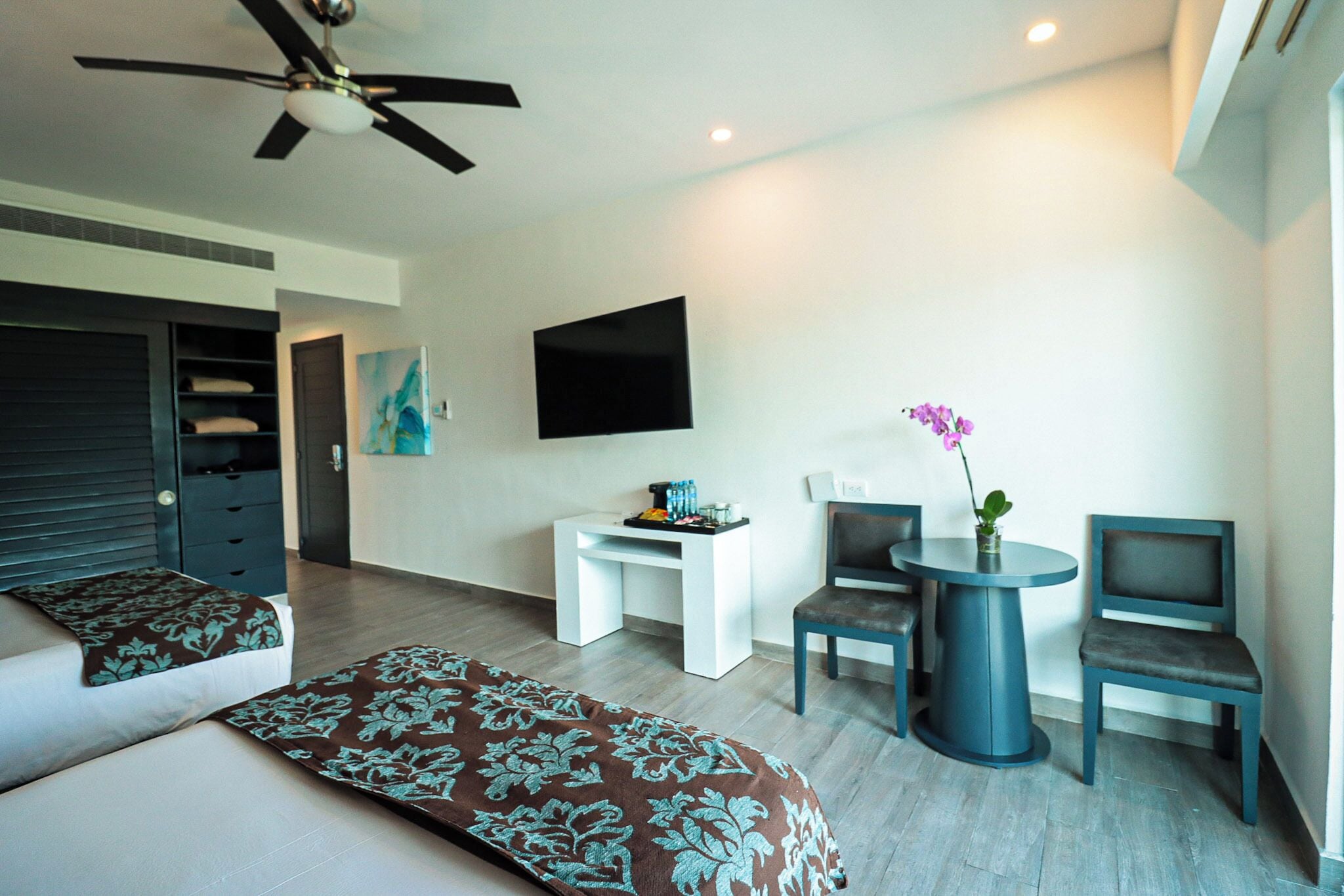 Een moderne hotelkamer met twee bedden, een plafondventilator, een flatscreen televisie, een kleintje tafeltje met een bloevas en twee stoelen.