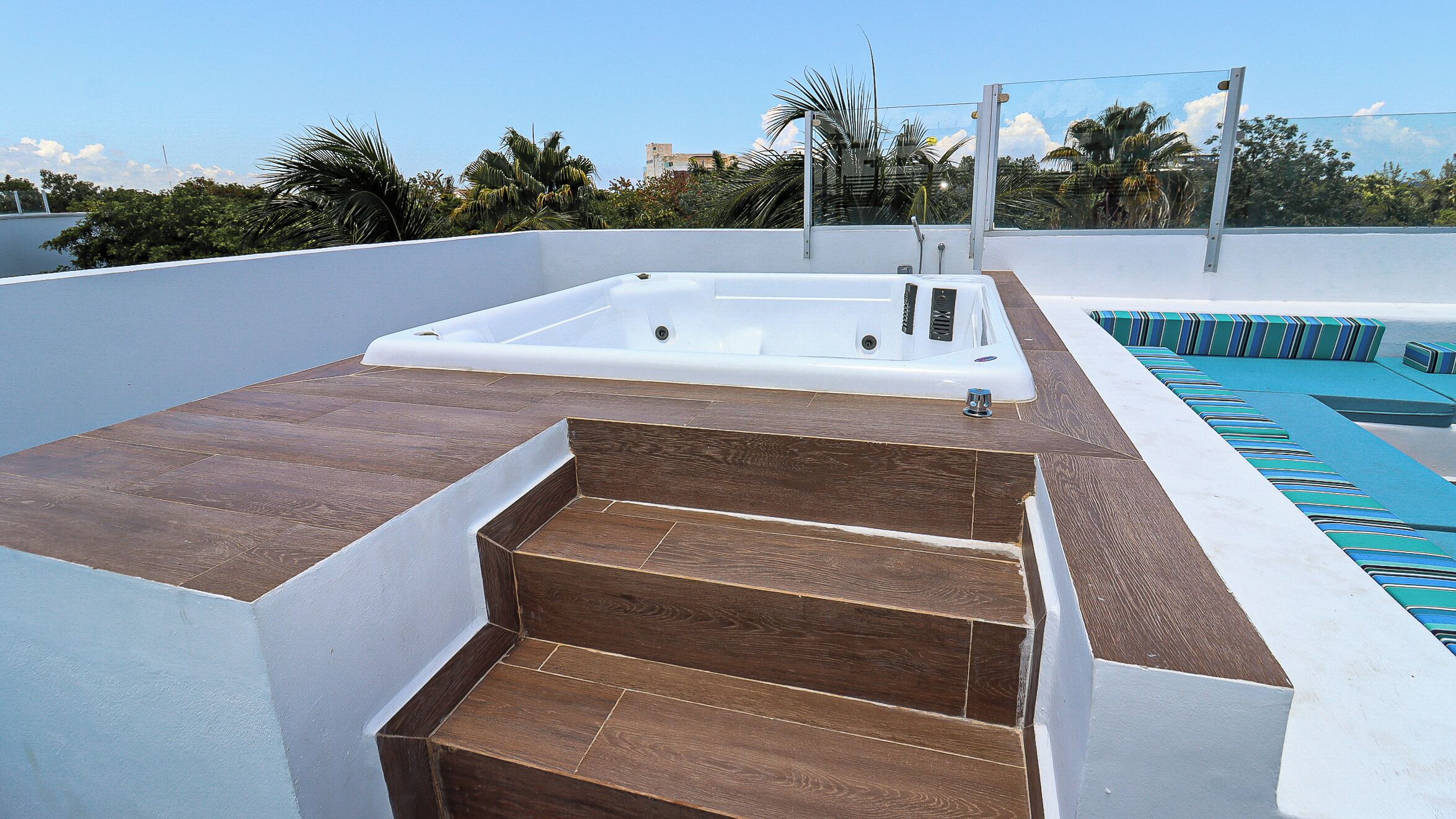 A rooftop pool area with a hot tub, wooden steps, and a pool with blue and green striped lounge area.
