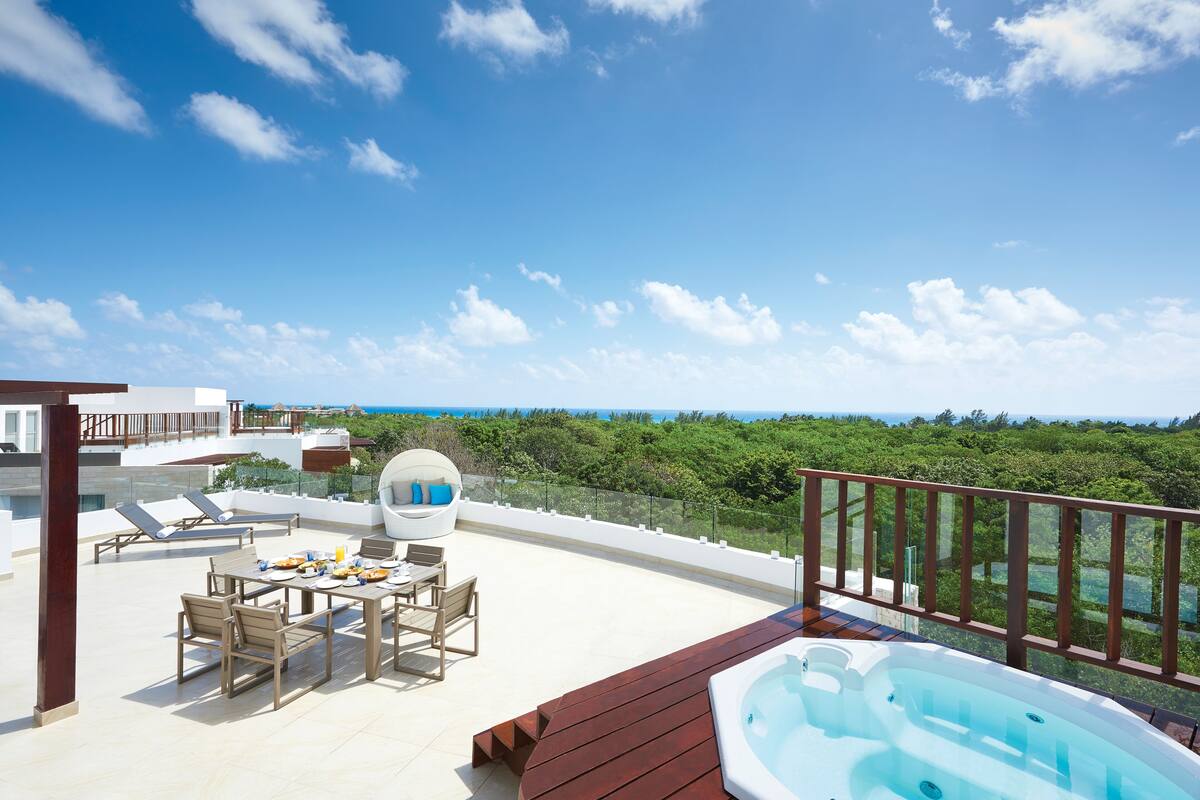 A rooftop terrace with a hot tub, lounge chairs, and a dining area overlooking a lush landscape.