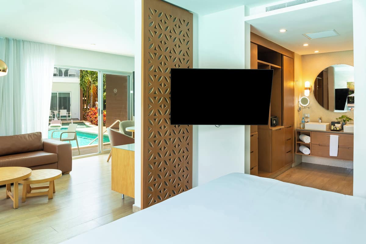 A modern hotel room with a TV, a sofa, a dining table, a kitchen area, and a view of a pool.