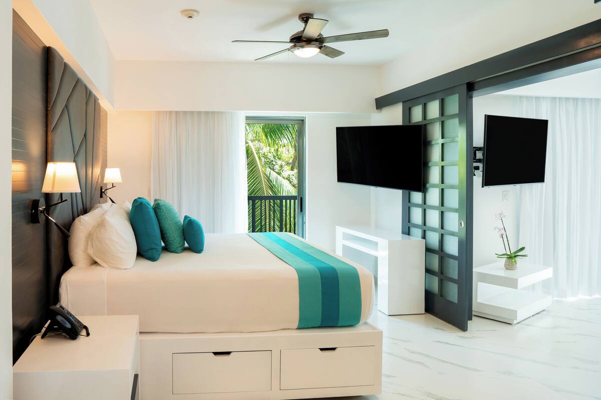 A modern hotel room with a large bed, a ceiling fan, a TV, and a sliding glass door to the outside.