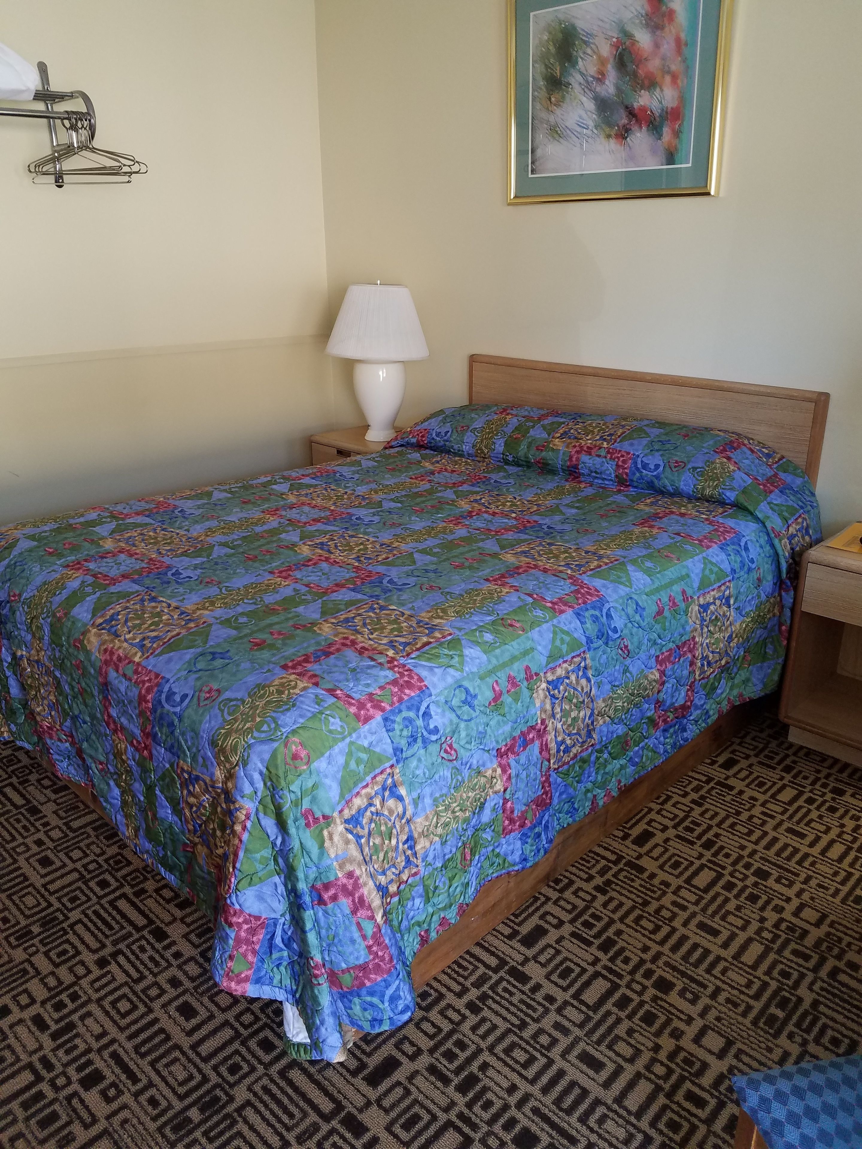 Una cama con un edredón colorido, una lámpara de noche, un cabecero de madera, una alfombra estampada y un cuadro en la pared.