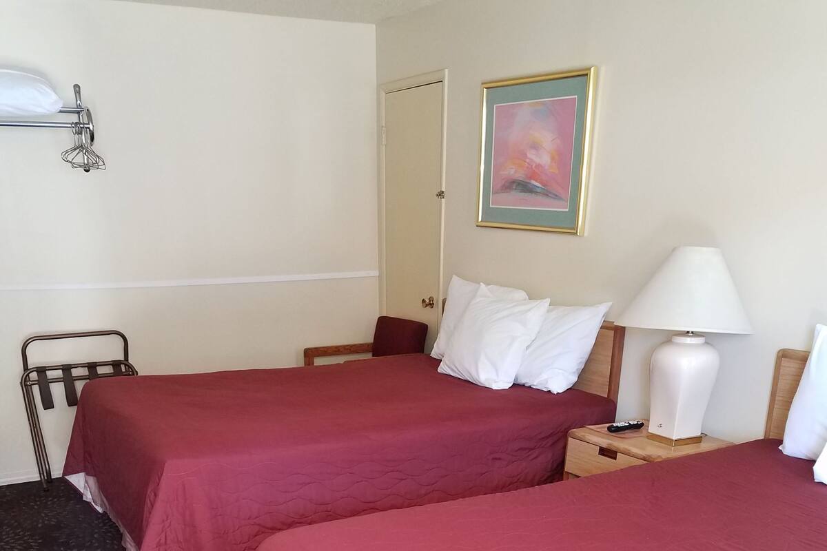 A hotel room with two beds, a nightstand with a lamp, a painting on the wall, and a door.