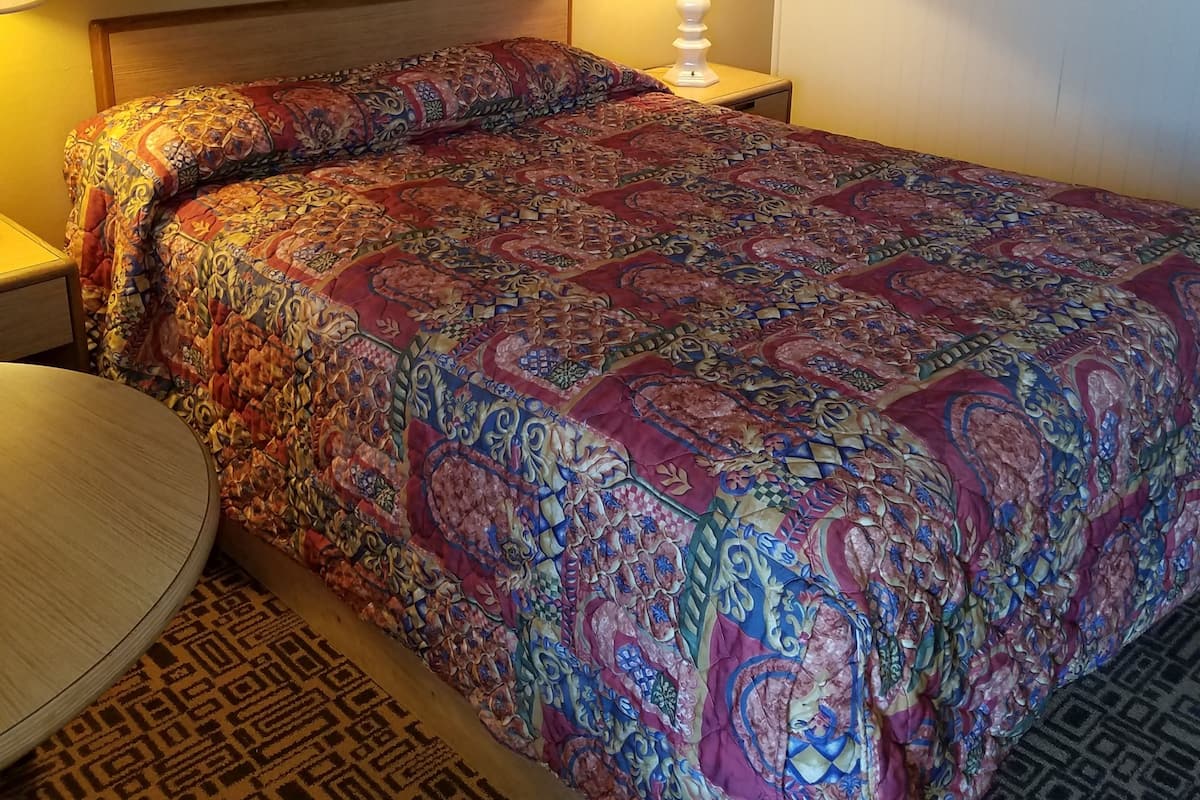 A hotel room with a large bed, a bedside table, a lamp, a framed picture on the wall, and a patterned rug.