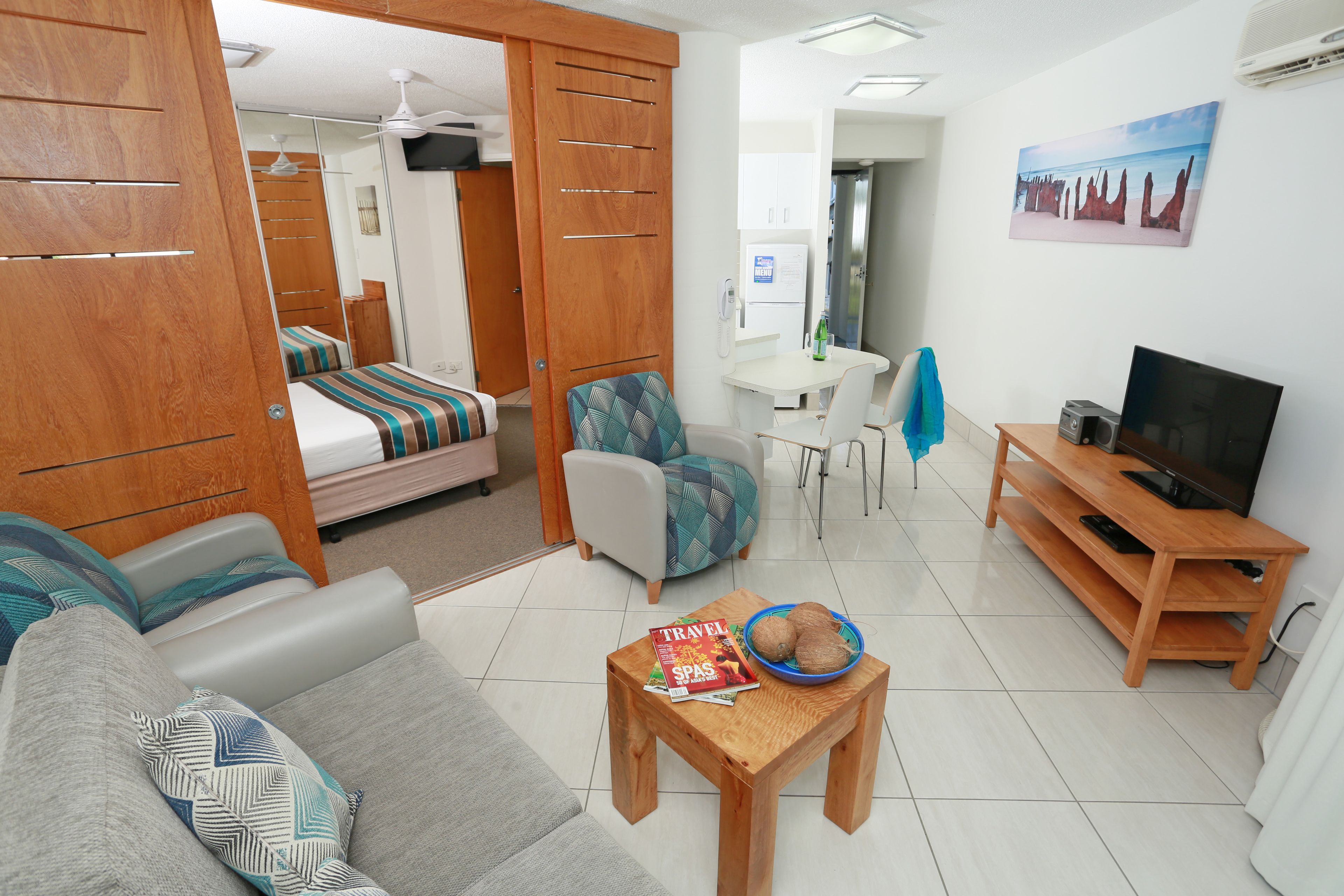 A compact hotel room with a bed, sofa, TV, dining table, and a small table with a magazine and a bowl of coconuts.