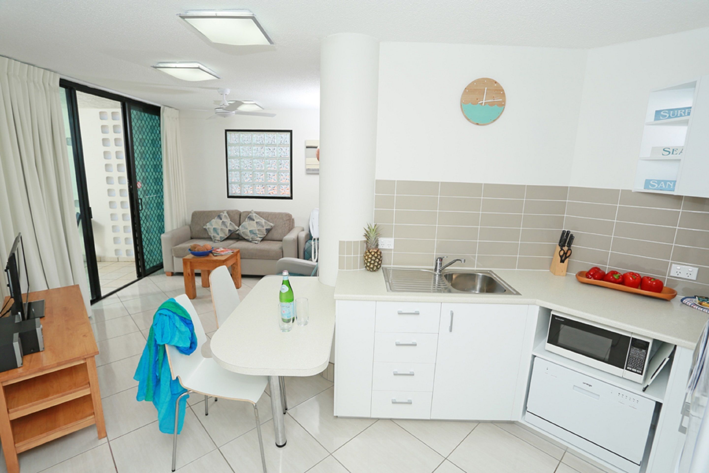 A modern kitchen with white cabinets, a dining table, and a small island. There is a clock on the wall and a window with a view of the outside.