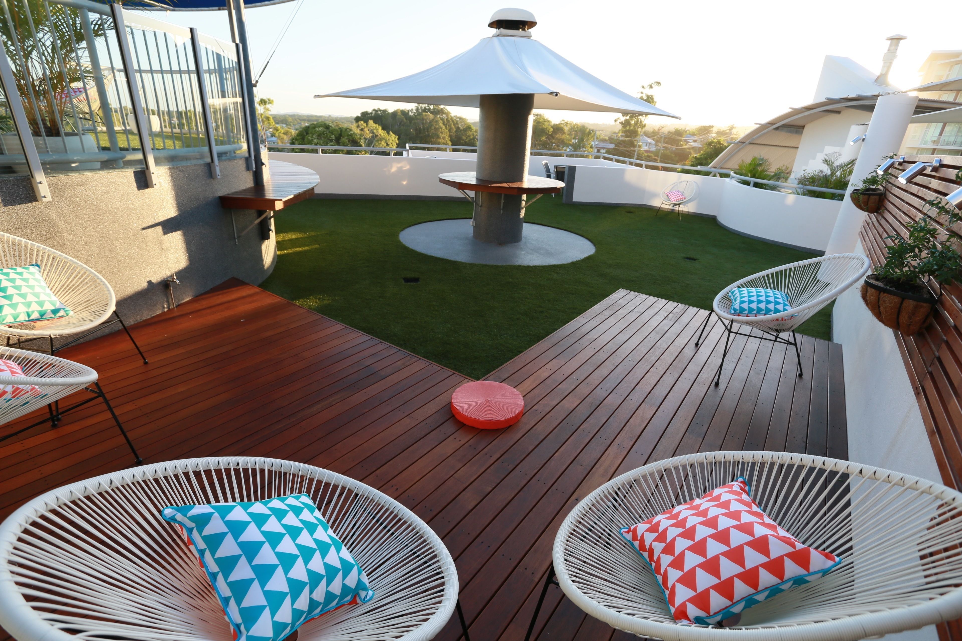 A rooftop patio with wooden flooring, white wicker chairs with patterned cushions, a central table with a white umbrella, and a small lawn area.