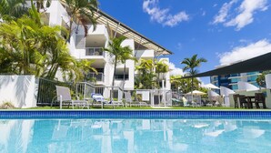 A swimming pool with lounge chairs and umbrellas, surrounded by palm trees and a multi-story building with balconies. - Beach Club Resort (Mooloolaba)