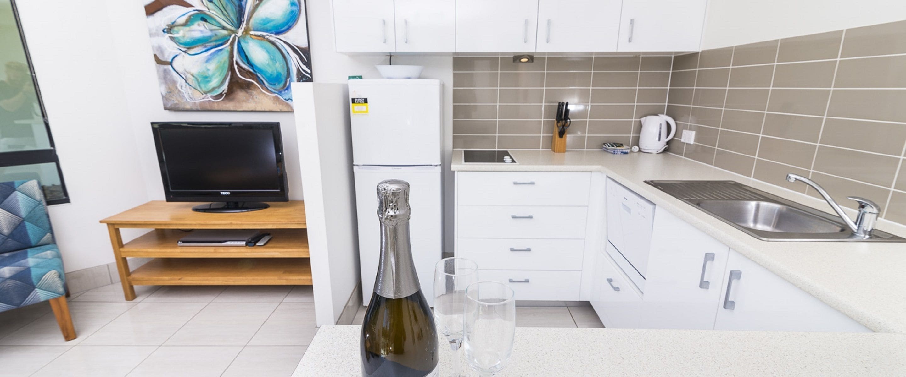 A modern kitchen with white cabinets, a built-in oven, a sink, and a countertop with a bottle and glasses.