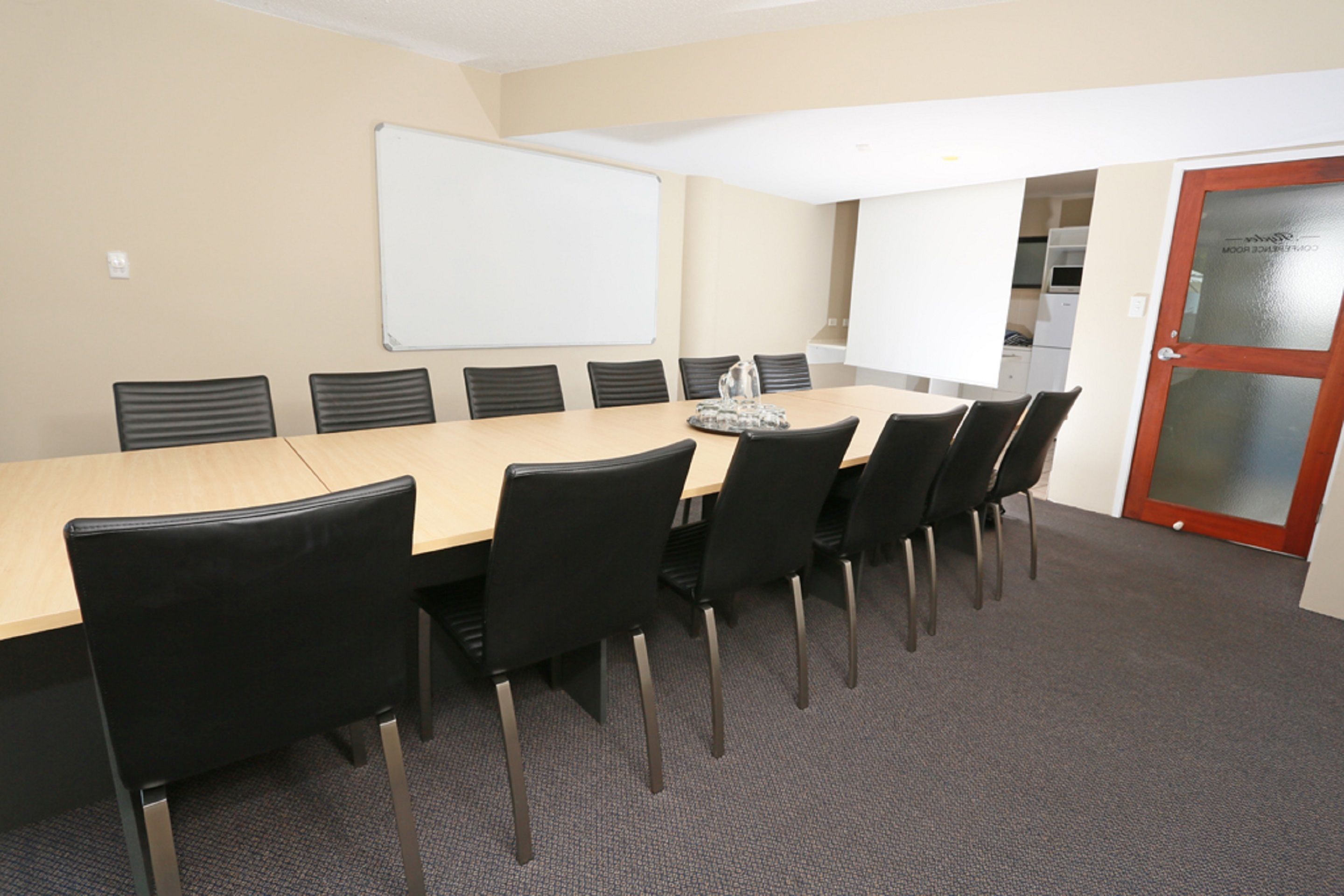 A conference room with a long table, black chairs, a whiteboard, and a door with a frosted glass panel.