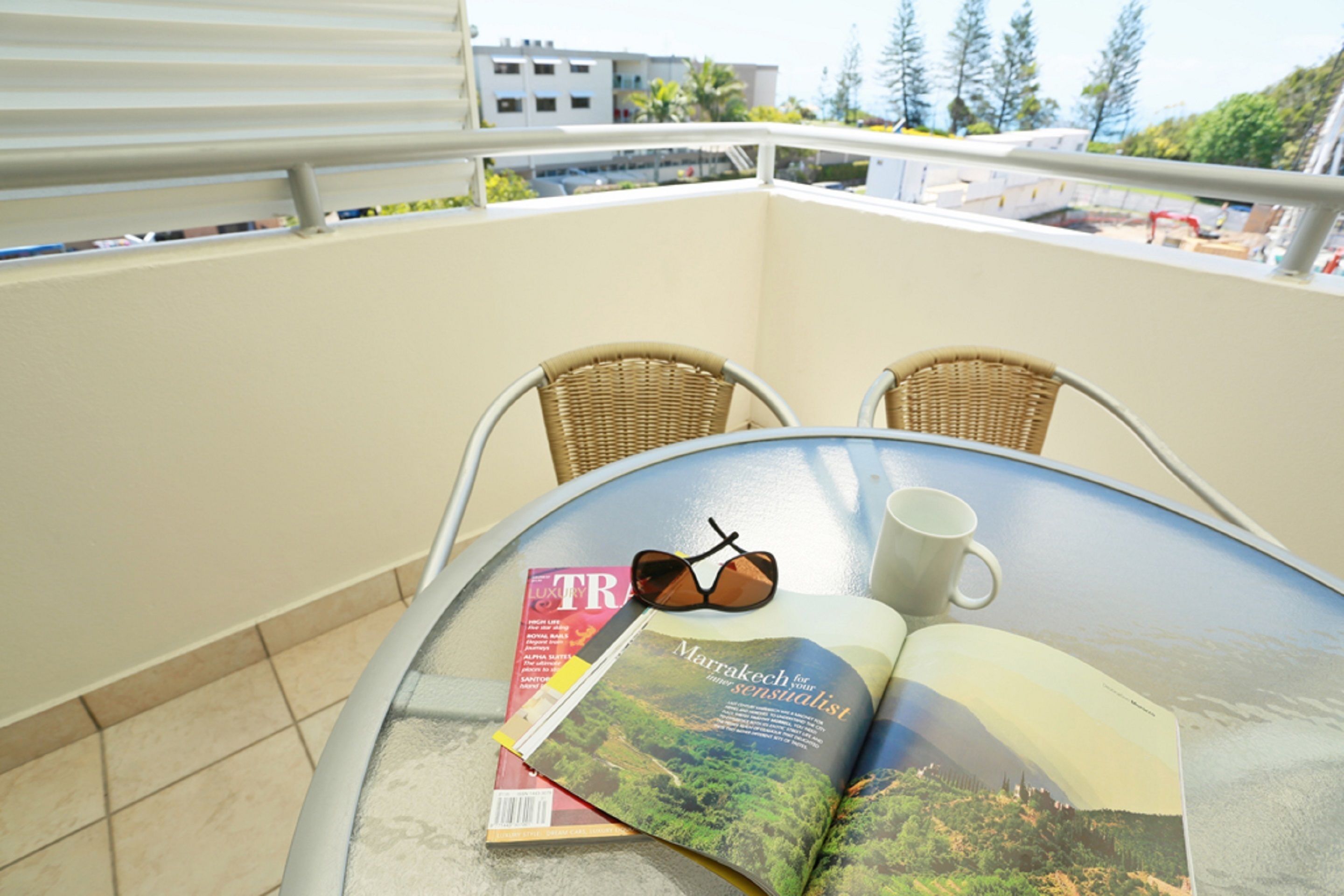A balcony with a table, two wicker chairs, a magazine, sunglasses, and a white mug.
