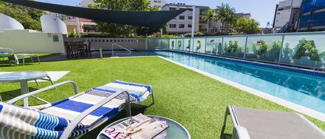 Un área de piscina en la azotea con sillones de descanso, una mesita y un separador de vidrio con vista a la ciudad.