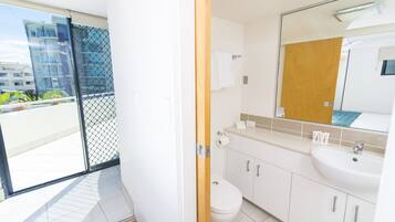Un baño moderno con inodoro, lavabo y espejo. Se ve un balcón con barandilla y edificios al fondo.