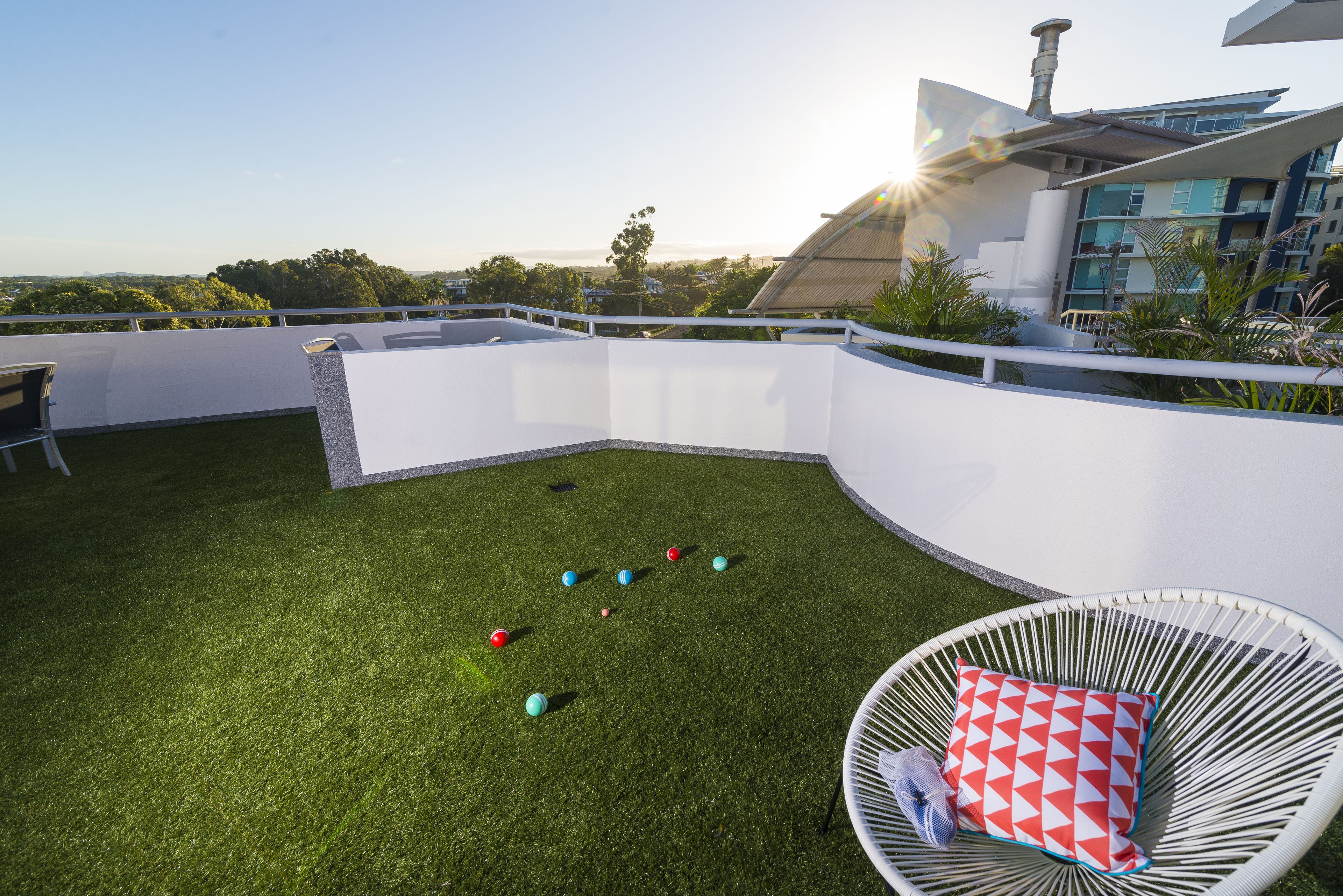Une terrasse sur le toit avec un mur blanc incurvé, une chaise blanche avec un coussin rouge et blanc, et une rangée de balles colorées sur l’herbe.