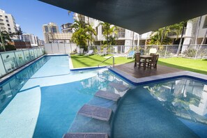 A swimming pool with steps leading into it, surrounded by a deck area with chairs and tables, and a view of city buildings in the background.