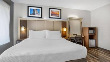 Quarto de hotel com uma cama grande, uma escrivaninha com um computador, um cesto de lixo pequeno e duas fotos emolduradas na parede.