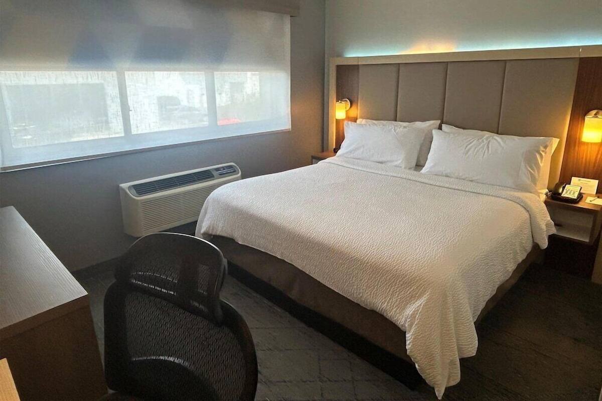 Une chambre d’hôtel comprenant un lit, un bureau, une chaise, une fenêtre avec des stores, un radiateur fixé au mur et une lampe de chevet.