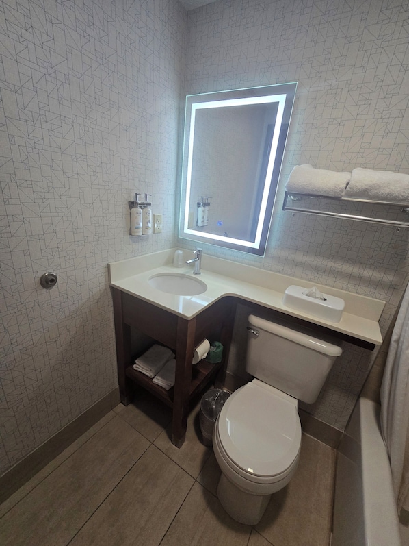 A modern bathroom with a geometric patterned wall, a large mirror with underglow lighting, a white sink, a toilet, and a towel rack.