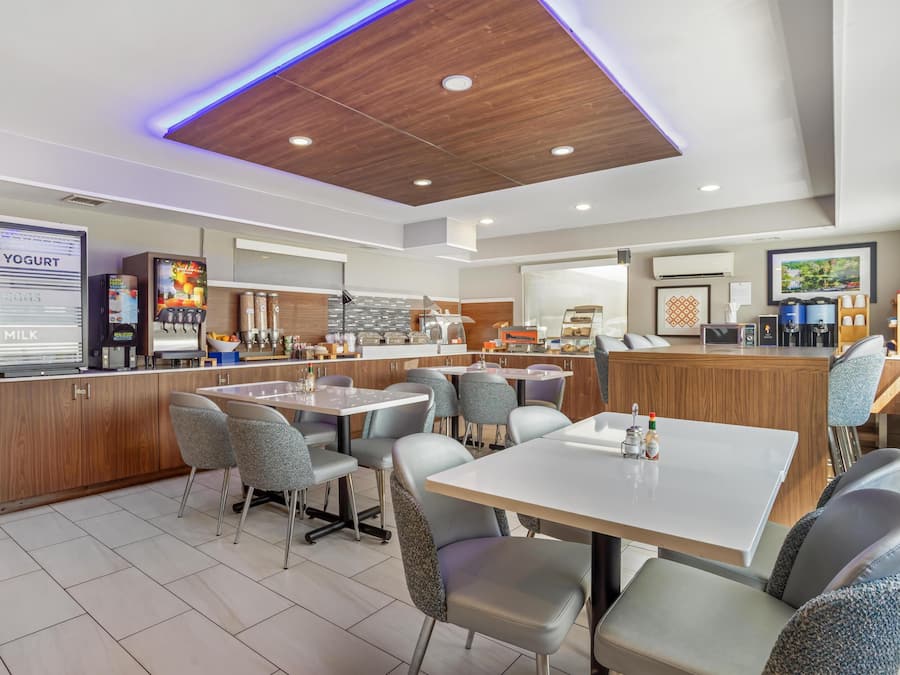 Uma área de refeições moderna com uma mesa central e cadeiras, uma máquina de iogurte e café, e um balcão com diversas opções de comida.