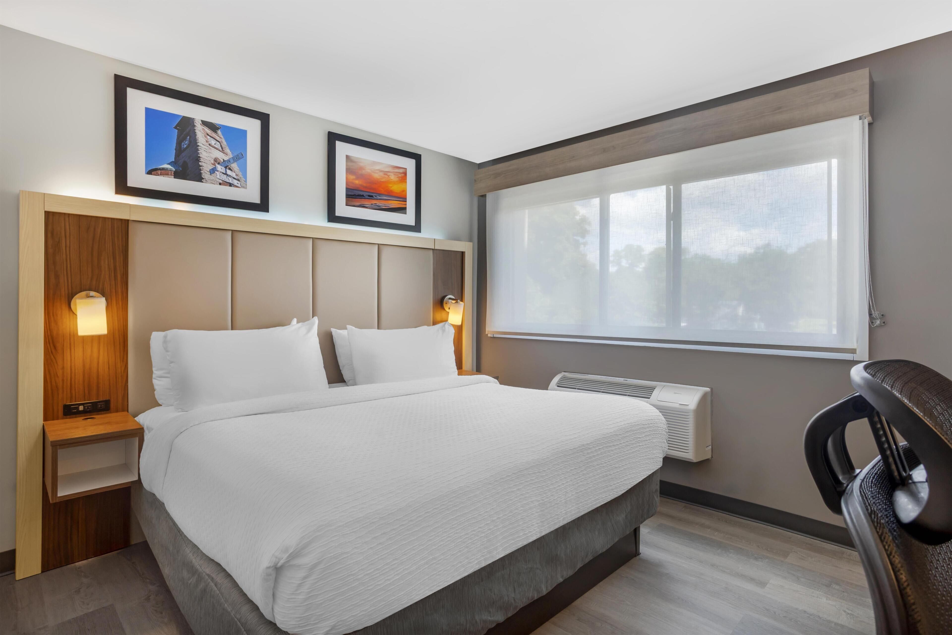 Habitación de hotel con cama, mesita de noche, escritorio, silla, ventana y dos cuadros en la pared.