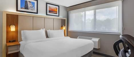 Habitación de hotel con cama, mesita de noche, escritorio, silla, ventana y dos cuadros en la pared.
