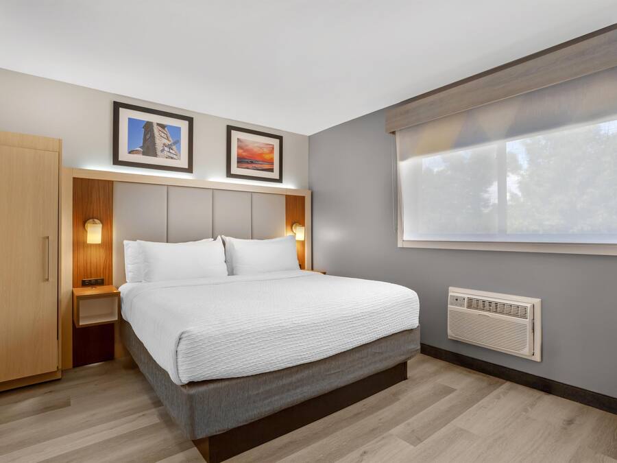 A modern hotel room with a bed, bedside tables, a wardrobe, a window with blinds, and two framed pictures on the wall.