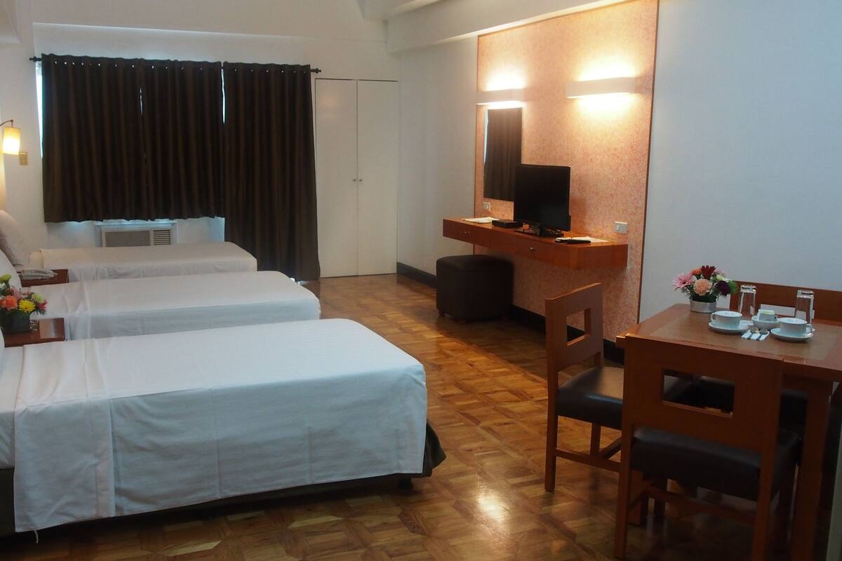 A hotel room with two beds, a desk, a chair, a TV, and a small table with a vase of flowers.
