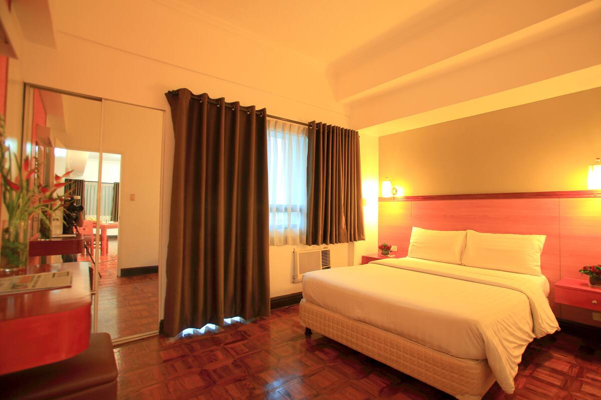 A hotel room with a bed, a nightstand, a lamp, a window with curtains, and a wooden floor.