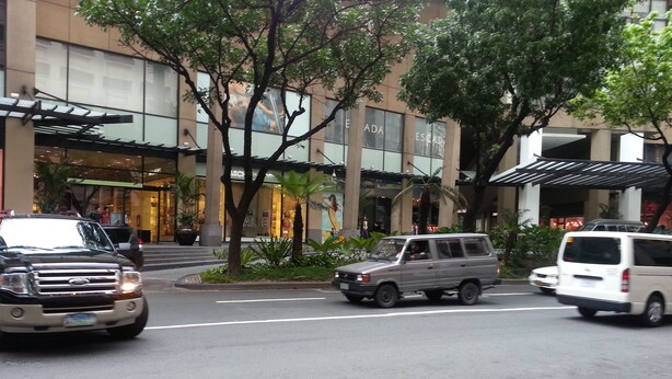 A street with a building featuring a modern facade, palm trees, and a pedestrian area with a covered walkway. - BSA Tower Serviced Residences (Makati)