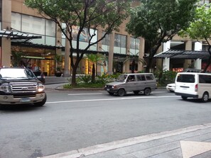 A street with a building featuring a modern facade, palm trees, and a pedestrian area with a covered walkway. - BSA Tower Serviced Residences (Makati)