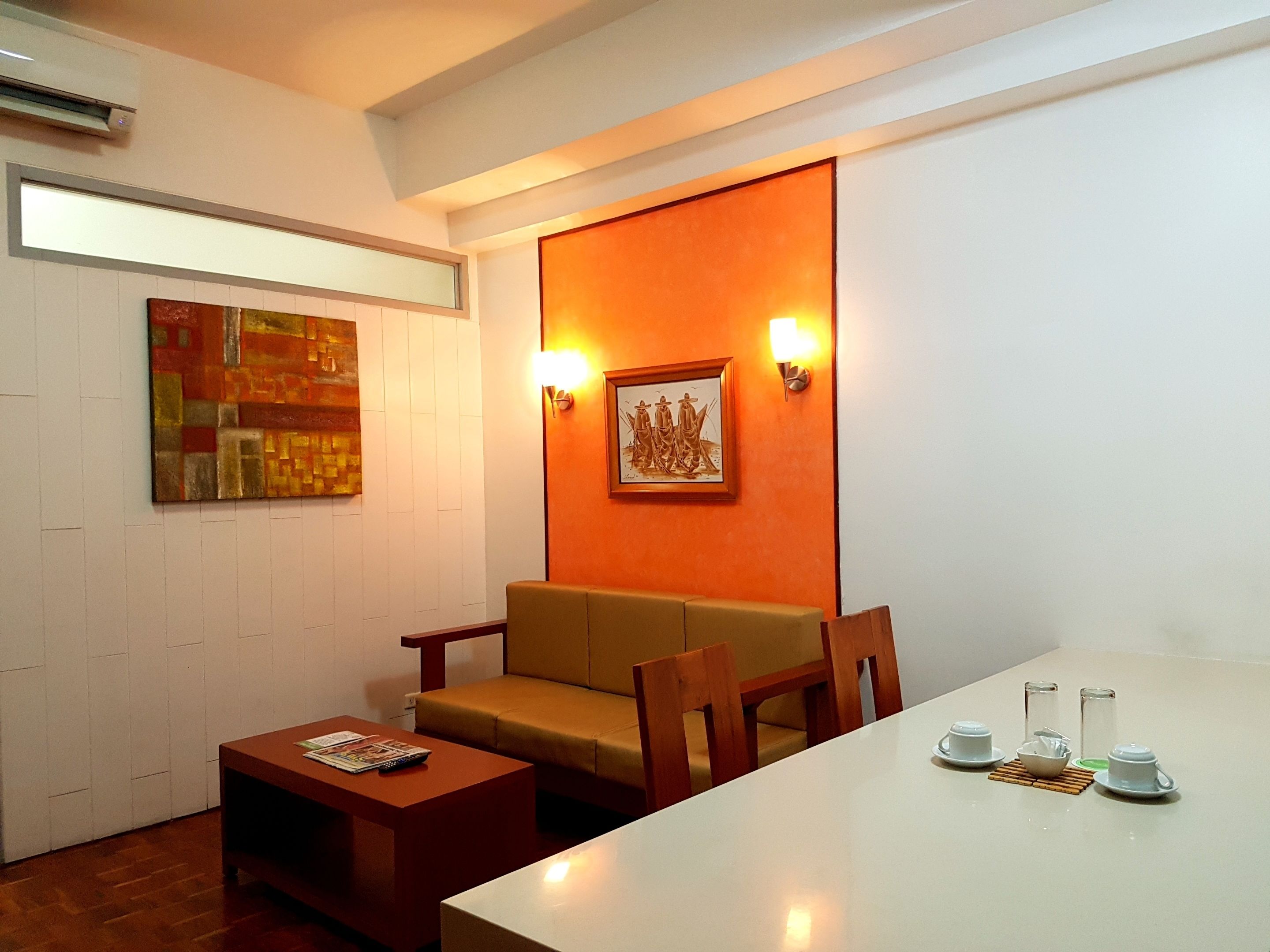 A modern hotel room with a white table, a brown sofa, and a small coffee table. There is a framed picture on the wall and a painting above the sofa.