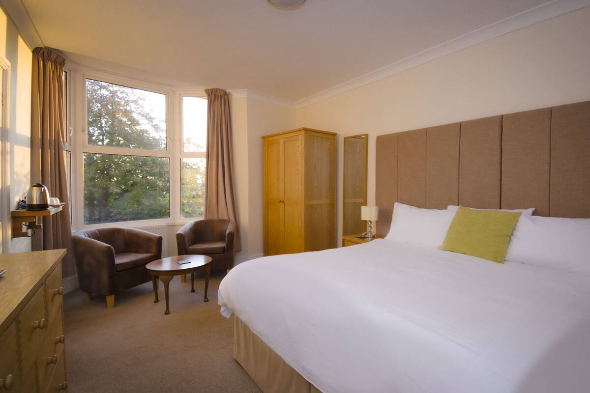 A hotel room with a large bed, a desk with a chair, a wardrobe, and a window with curtains.