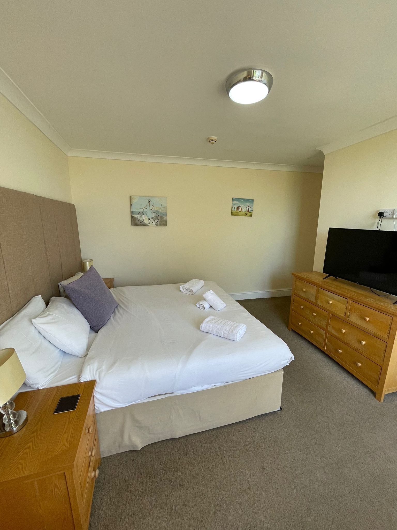 A hotel room with a bed, a wooden dresser, a television, and two wall art pieces.