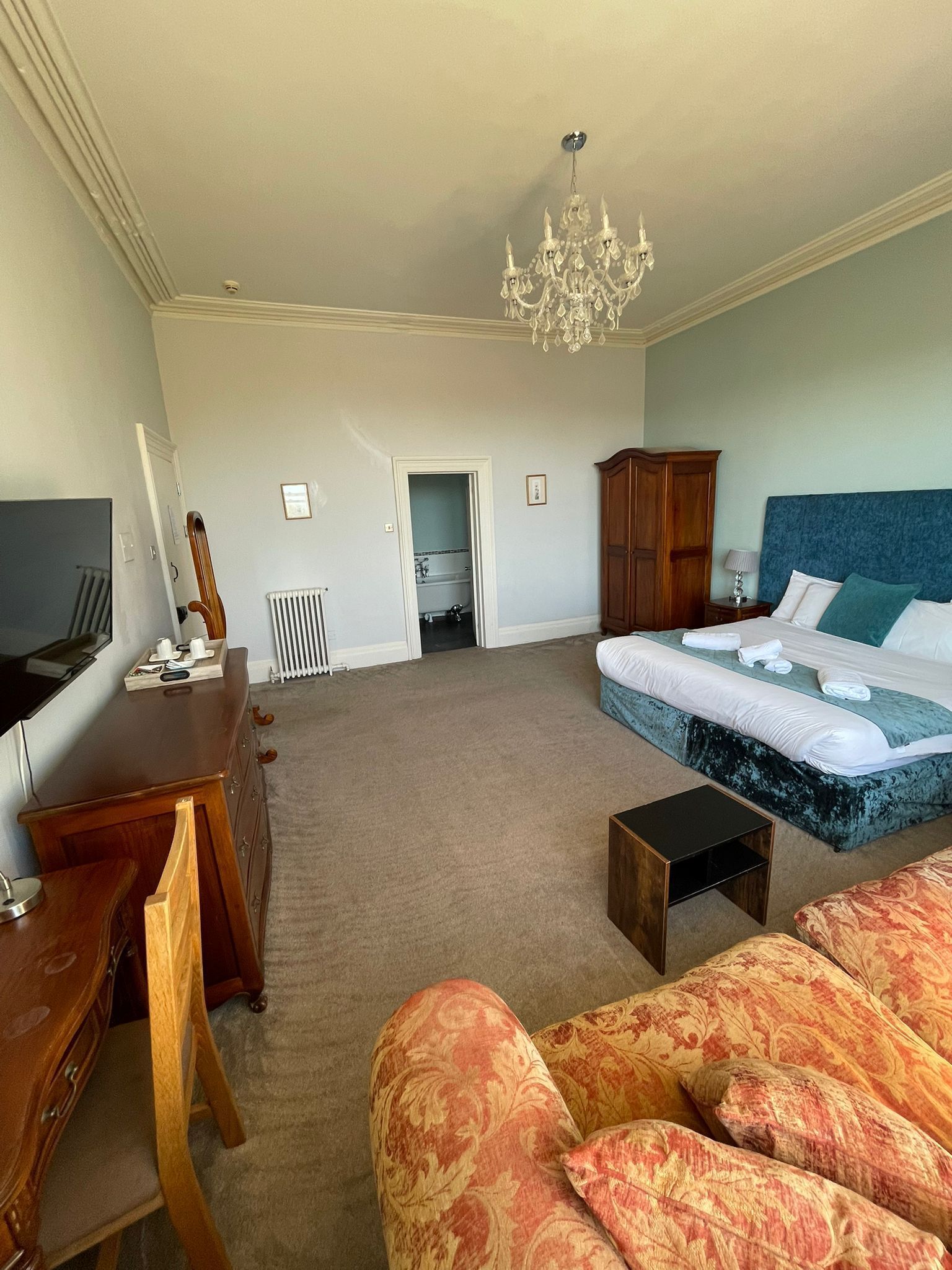 A spacious hotel room with a large bed, a desk, a television, and a chandelier.