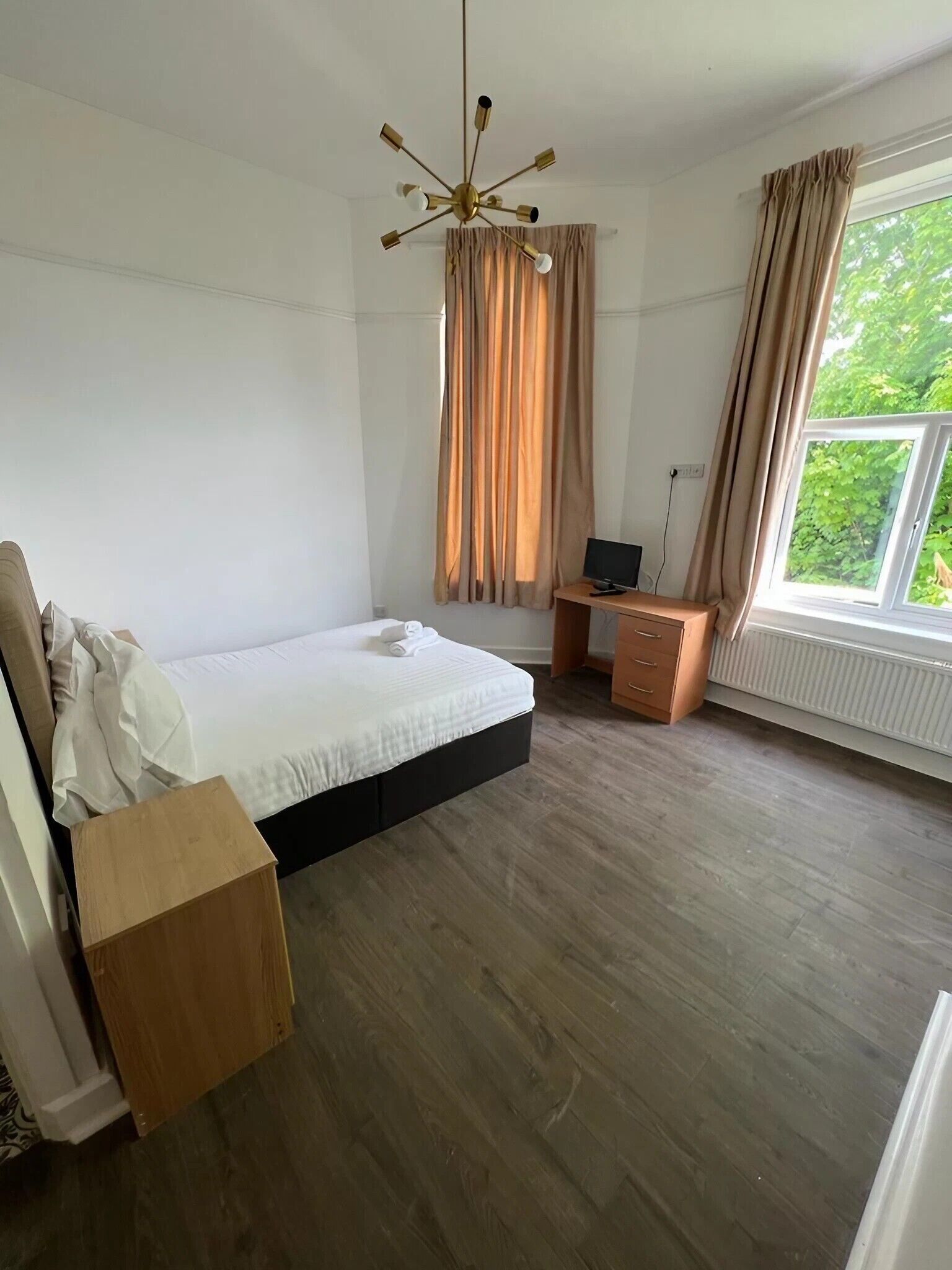 Une chambre à coucher avec un lit, un bureau, une chaise, un radiateur et une fenêtre avec des rideaux.