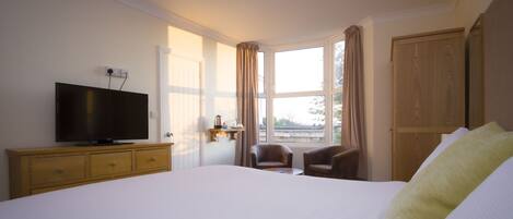 Habitación de hotel con una cama grande, un televisor en una cómoda de madera, dos sillas, una ventana con cortinas y vistas al exterior.