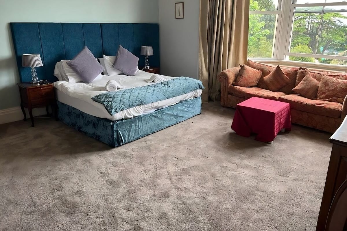 A bedroom with a bed, a nightstand, a red table, a brown sofa, and a window with curtains.