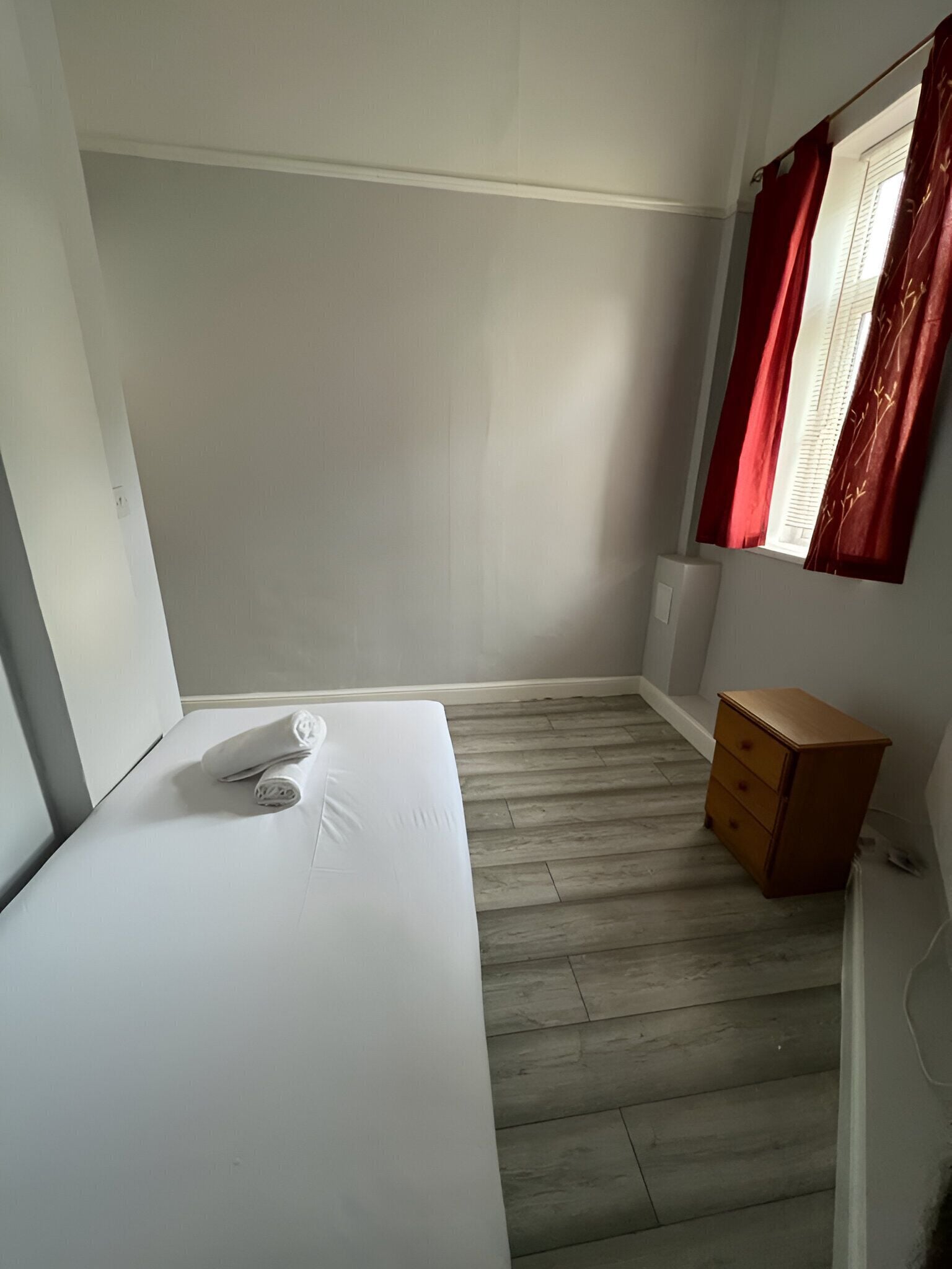 A small, sparsely furnished room with a single bed, a wooden dresser, and a window with red curtains.