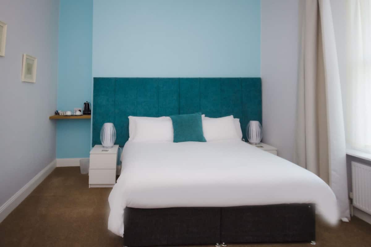 A bedroom with a large bed, a nightstand, a shelf with items, and a window with curtains.