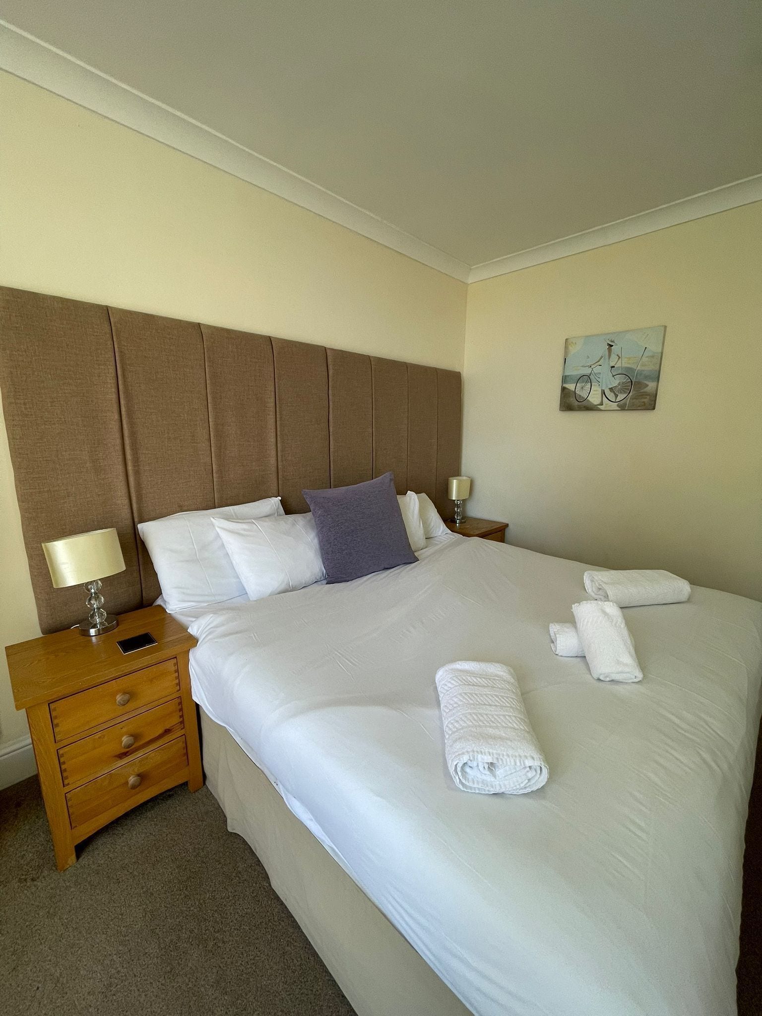 A neatly made bed with white linens, a wooden nightstand with a lamp, and a small painting on the wall.