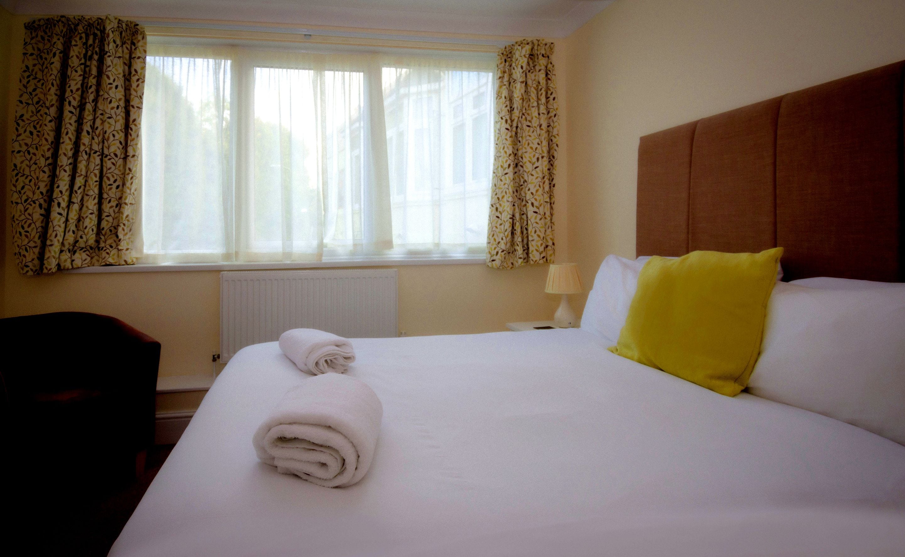 Une chambre d’hôtel avec un grand lit, une tête de lit, une fenêtre avec des rideaux, un radiateur et une chaise.
