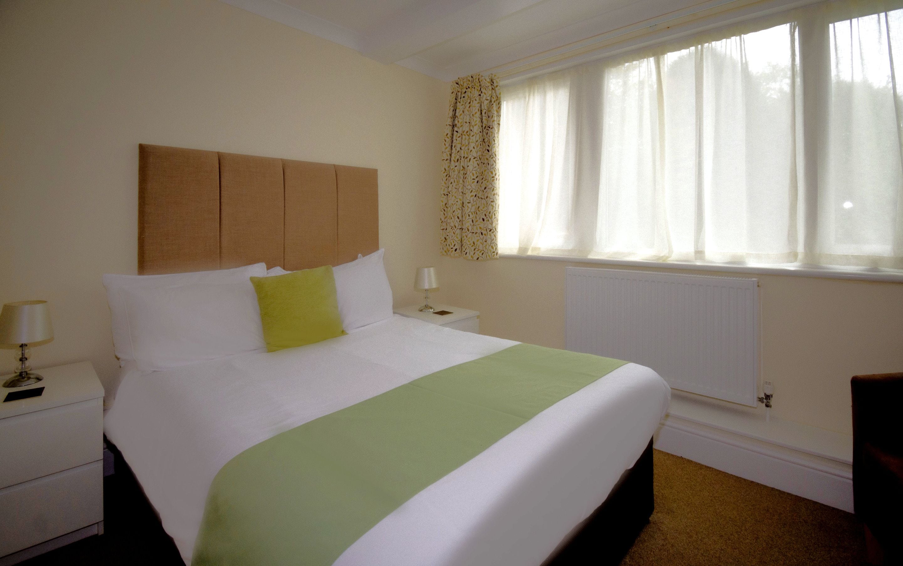 A hotel room with a bed, nightstand, lamp, and window with curtains.