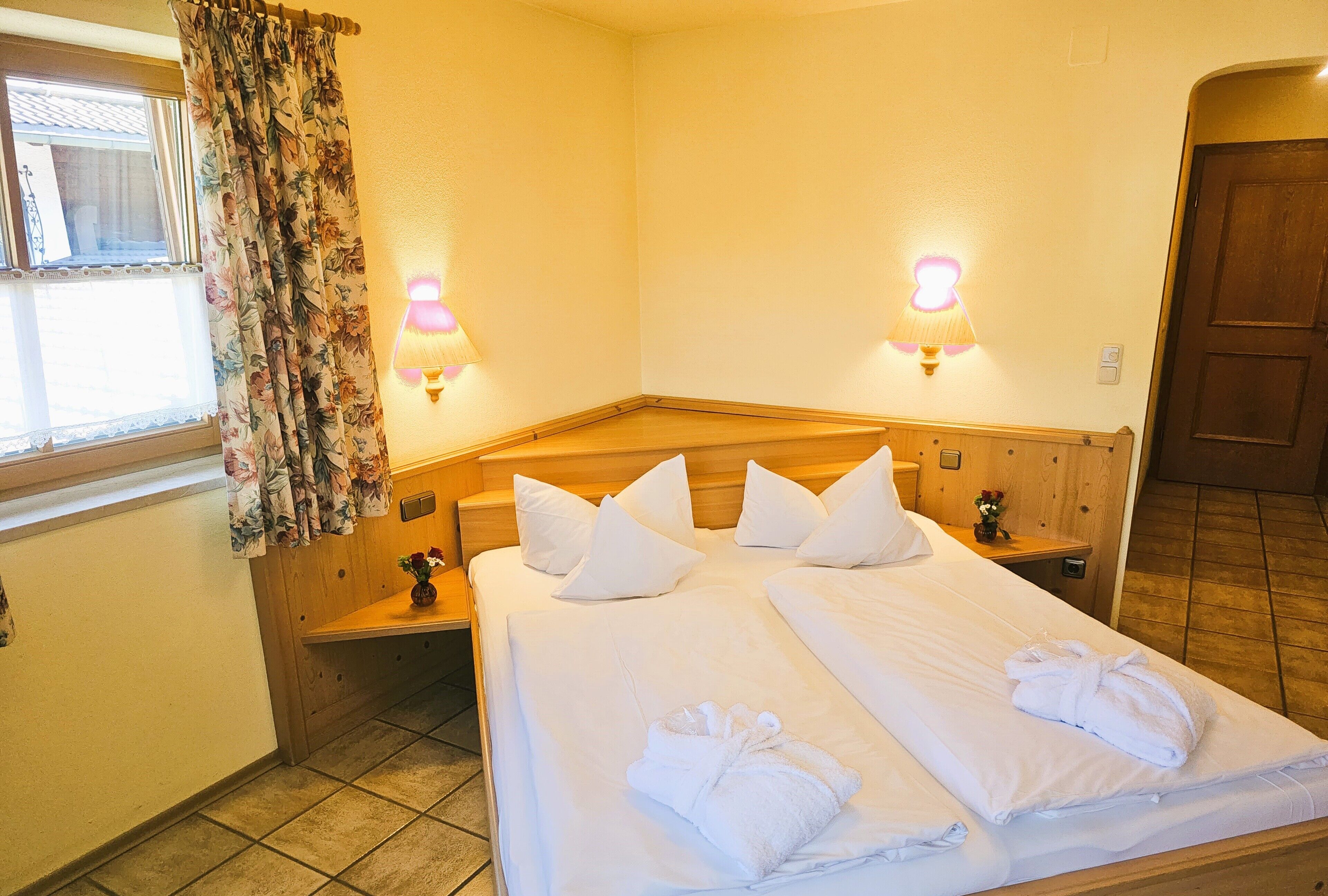 A hotel room with a large bed, two bedside tables, a wooden headboard, a window with floral curtains, and two wall-mounted lamps.