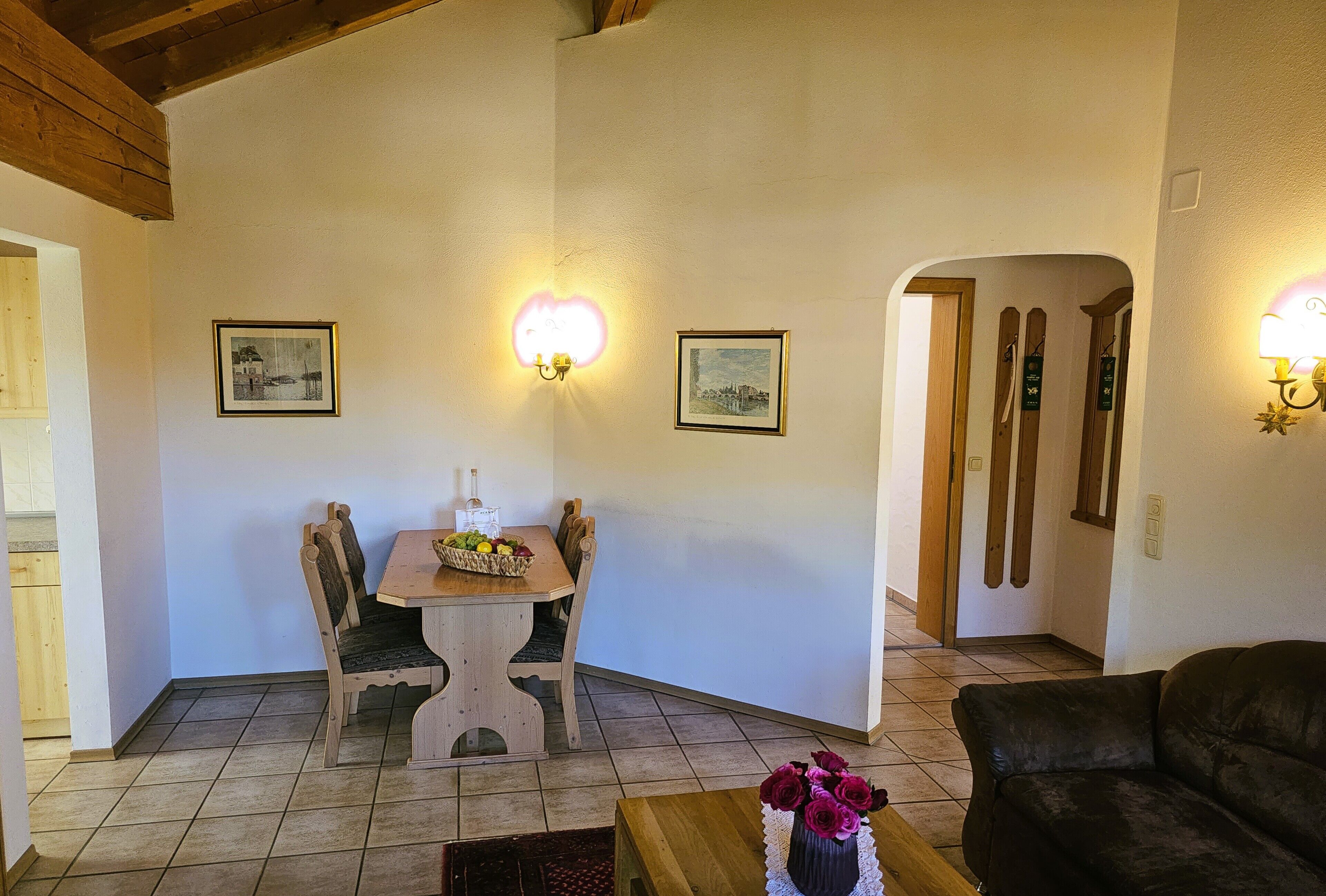 Un comedor con una mesa y sillas de madera, un florero con flores sobre una mesita auxiliar, y dos lámparas fijadas a la pared.